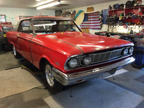 1964 Ford Fairlane - A red vintage car is parked in a garage, showing its front grille and headlights.
