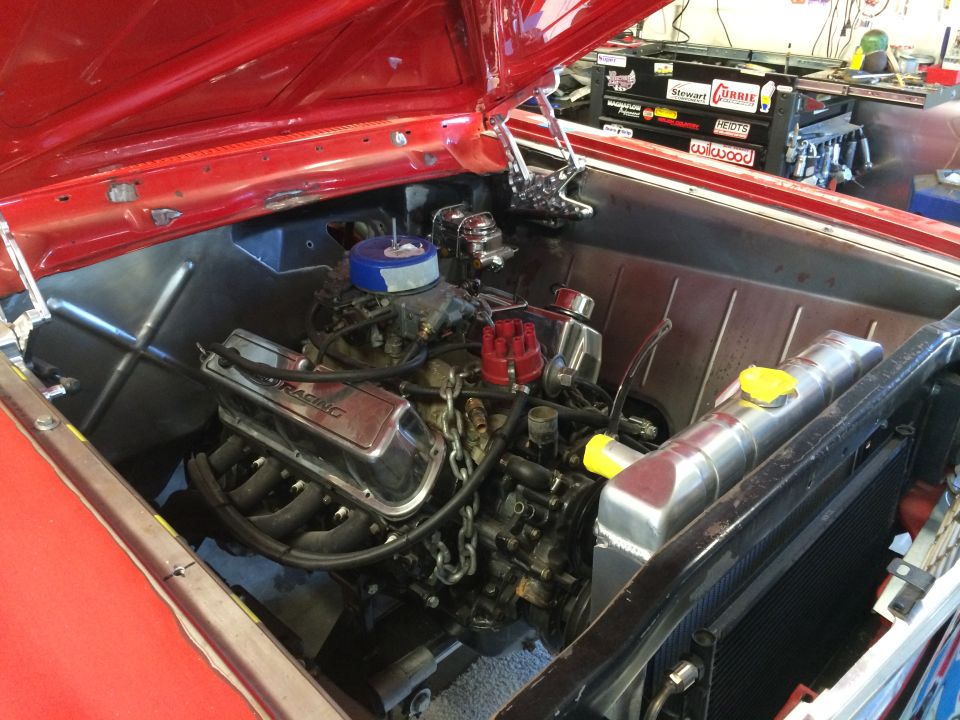 Red car engine bay with chrome valve covers and a blue air filter. A toolbox with stickers is visible in the background