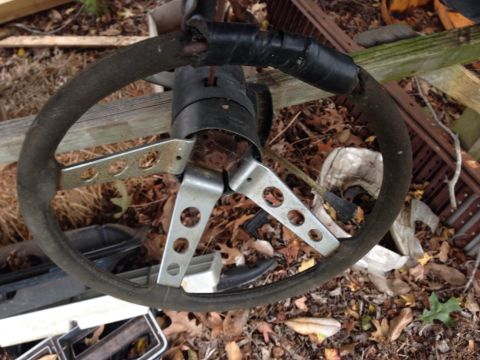1979 Ford Bronco - A weathered steering wheel with a black rim and silver spokes lies amidst fallen leaves and debris.