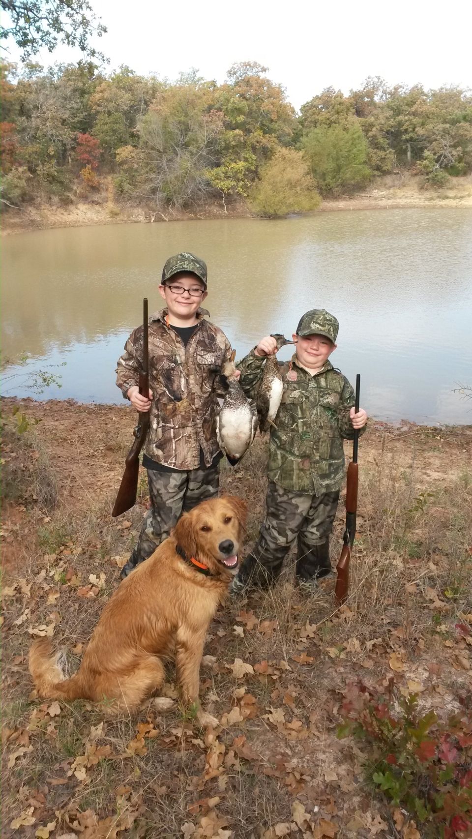 Two boys in camouflage with hunting rifles and a dog pose by a lake with their duck catch