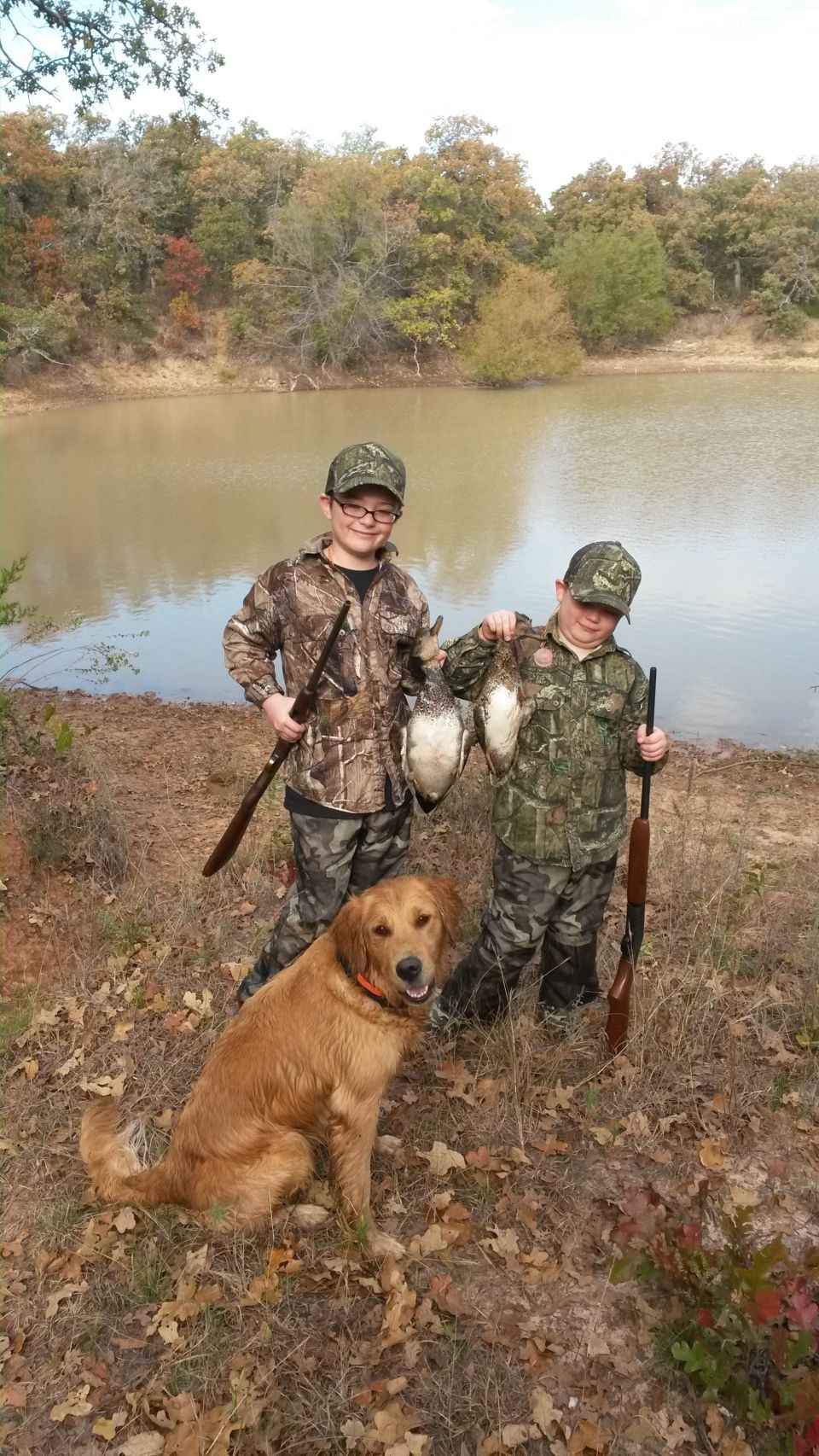 Two boys with hunting gear and ducks, a golden retriever, and a lake