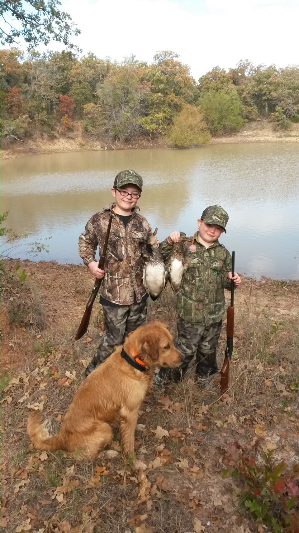 Two kids in camo with hunting rifles and ducks, a golden retriever sits nearby by a pond
