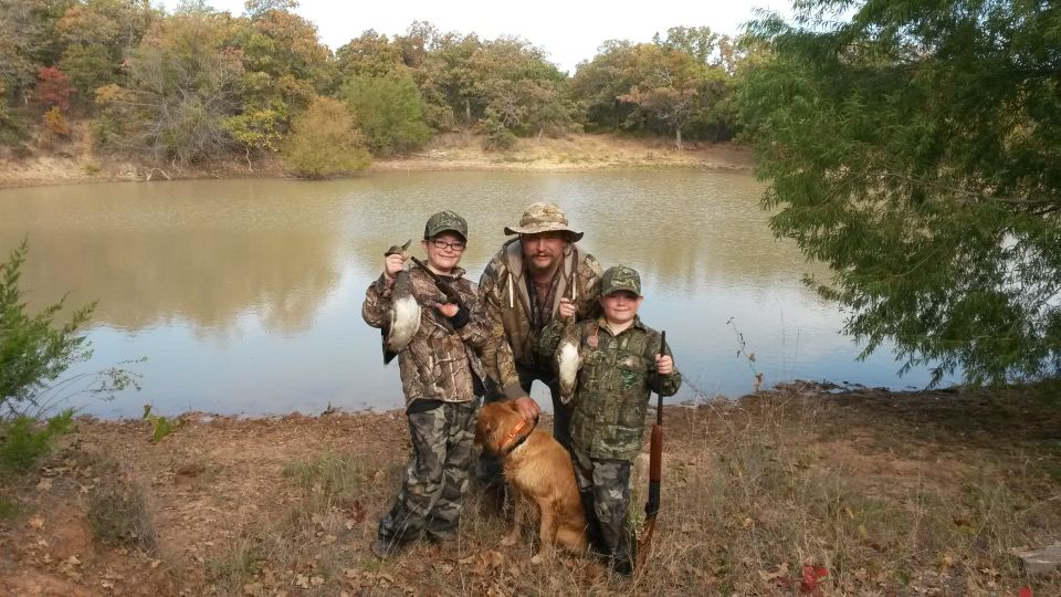 Man, two kids, and a dog pose by a lake, holding ducks they hunted