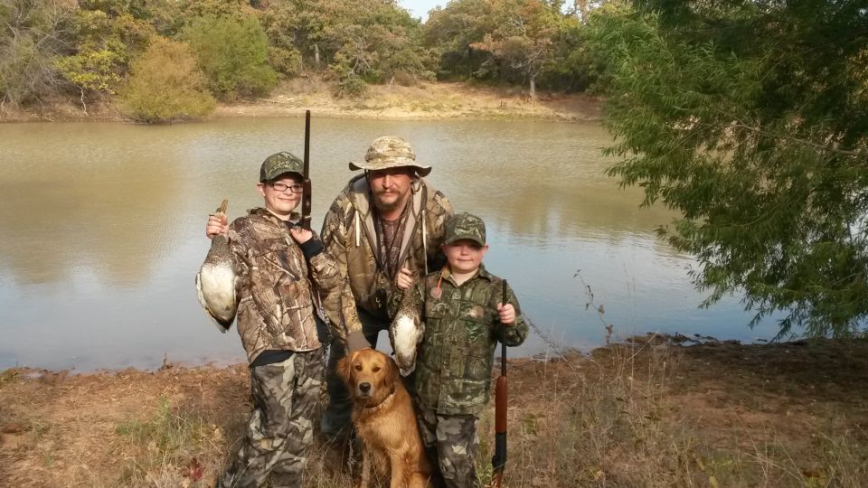 Three hunters in camouflage, a golden retriever, and two ducks stand by a lake