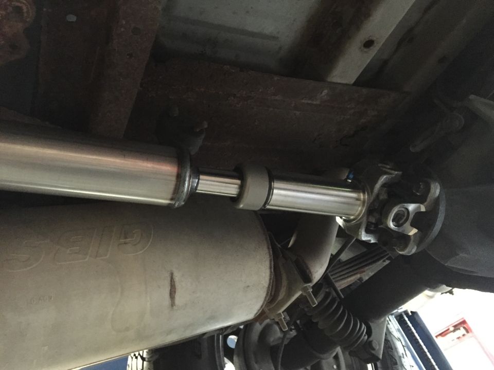 Underside view of a vehicle's driveshaft and exhaust system, showing a new muffler and U-joint