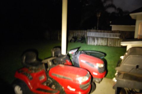 1994 Ford Bronco - Two red riding lawn tractors are parked on a grassy lawn next to a house at night. A ladder is leaning against a wooden fence in the backgr…