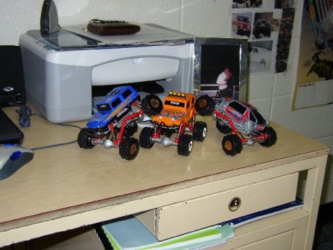 1989 Ford Bronco - Three remote-controlled monster trucks are lined up on a desk next to a printer and a framed photo.