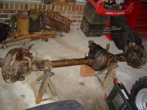 1986 Ford Bronco - A rusty front axle assembly is displayed on jack stands, with other vehicle parts scattered around.