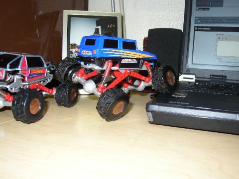 1989 Ford Bronco - Two remote control monster trucks are on a desk next to a laptop computer.
