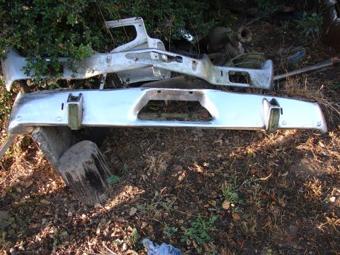 1973 Ford Ranchero - Discarded chrome bumper and associated frame parts lie on the ground amongst debris and foliage.