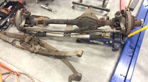1984 Ford Bronco - Front axle and leaf springs removed from a vehicle, resting on a shop floor.