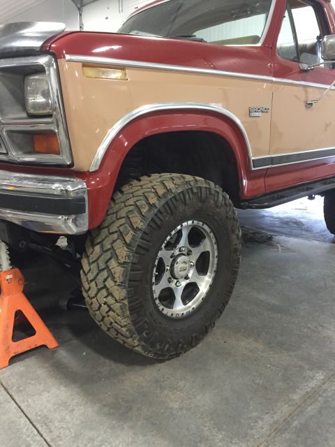 1984 Ford Bronco - A two-tone red and tan Ford Bronco is shown with its front wheel elevated by a jack, revealing aftermarket wheels and tires.
