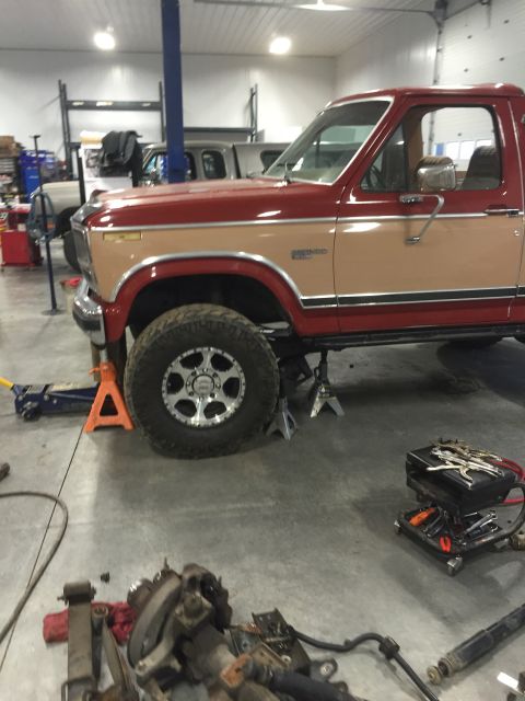 1984 Ford Bronco - A two-tone red and tan pickup truck is elevated on jack stands in a workshop, with another vehicle visible in the background.