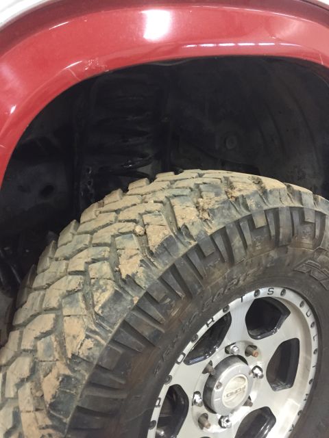 1984 Ford Bronco - Close-up of a red fender and a muddy off-road tire with a custom wheel, showing part of the suspension system.