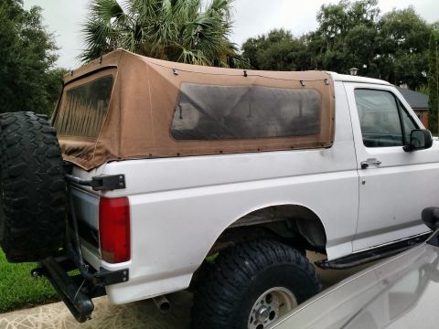 1992 Ford Bronco - Side rear view of a white lifted full-size Bronco fitted with a tan canvas soft top, swing-out spare tire carrier and mud tires.