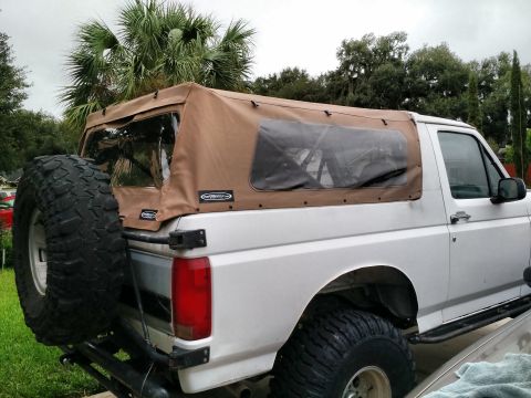 1992 Ford Bronco - silver Ford Bronco fitted with a tan aftermarket soft top, oversized mud tires and a rear-mounted spare sits in a yard.