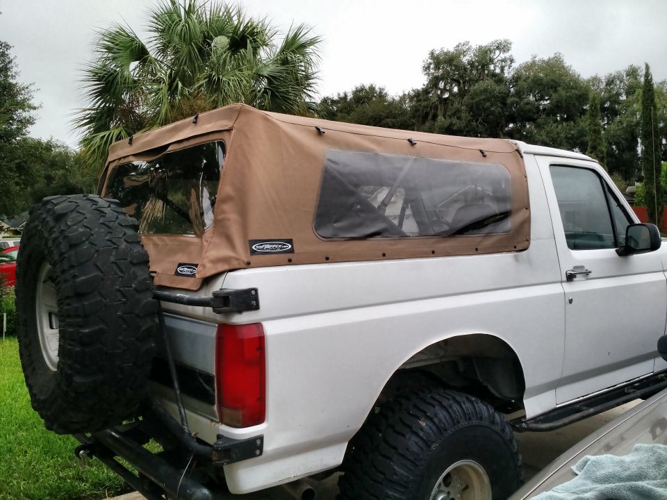 Silver Ford Bronco with tan soft top and large mud tires parked in a yard