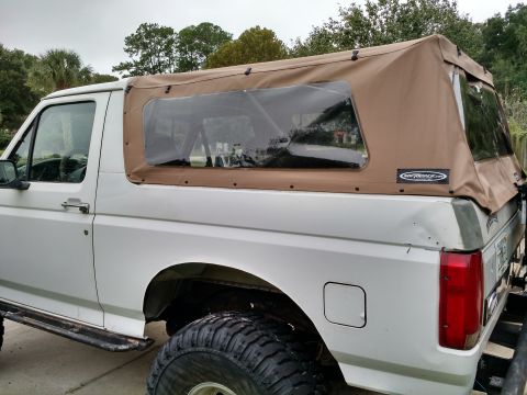 1992 Ford Bronco - White lifted full-size Ford Bronco fitted with a tan canvas soft top and clear rear window, on oversized tires outdoors.