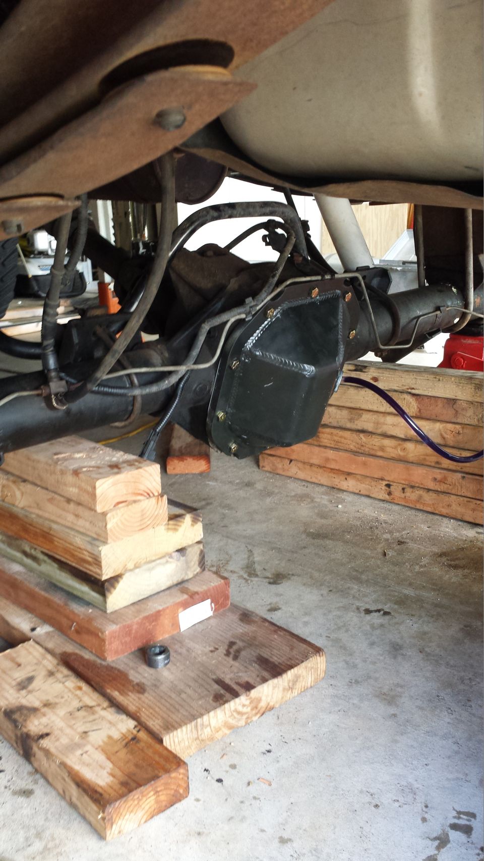 Rear differential assembly with black cover, supported by wooden blocks. Suspension components and fuel tank visible above