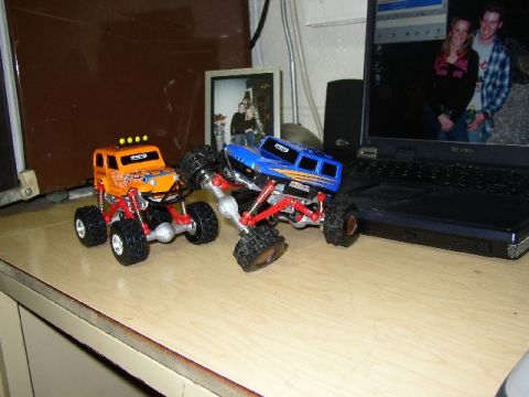 1989 Ford Bronco - Two remote-controlled monster trucks are positioned on a wooden desk surface.