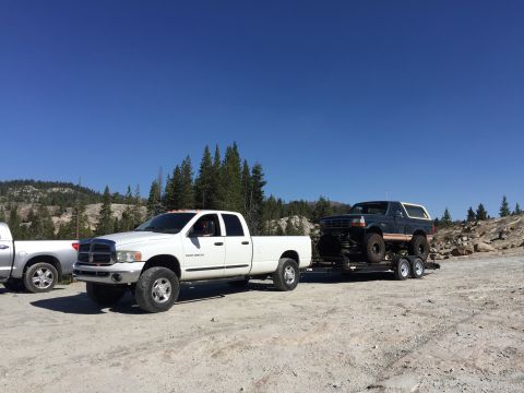 1989 Ford Bronco - A white pickup truck tows a lifted Ford Bronco on a trailer through a rocky, wooded area under a clear blue sky.