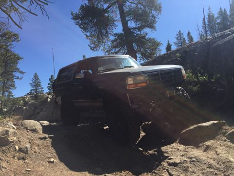 1989 Ford Bronco - A dark SUV navigates a challenging rocky trail under a clear blue sky.