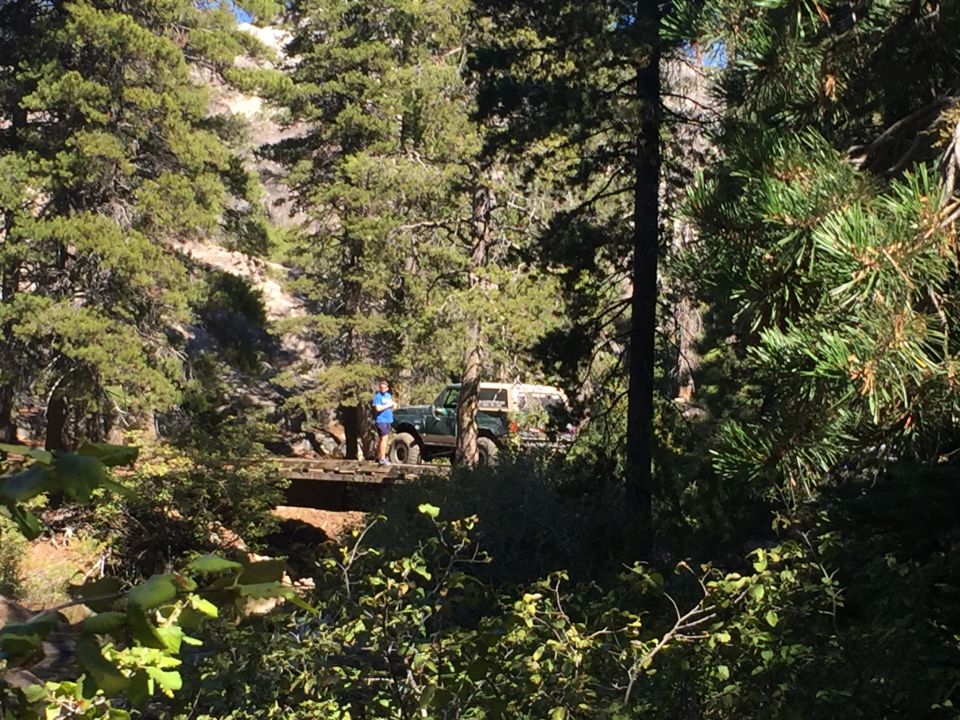 Green SUV with a white camper shell parked on a wooden bridge, two people standing nearby, surrounded by trees