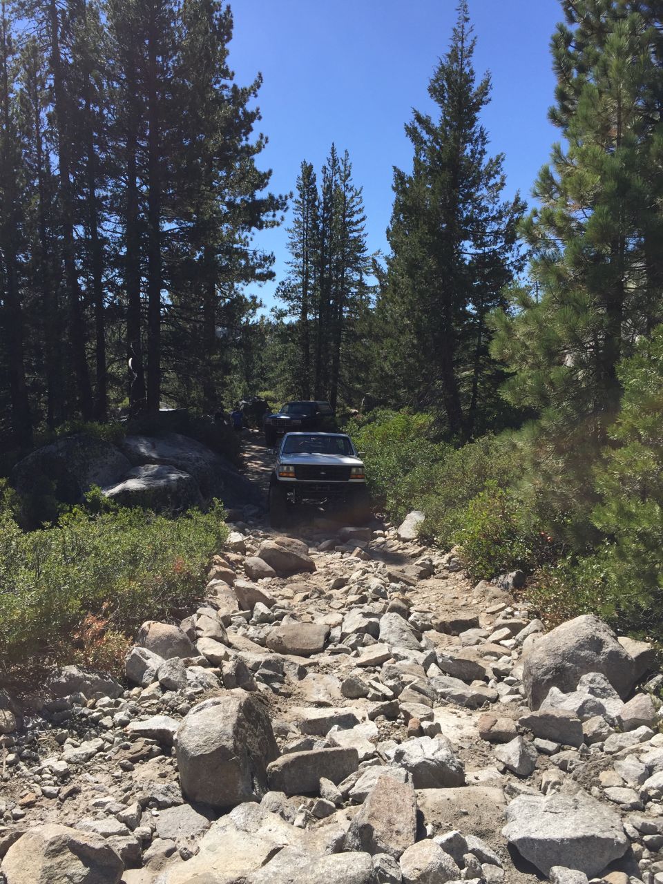 Silver 1987 Ford Bronco driving on a rocky trail, front-right quarter view, with trees and blue sky