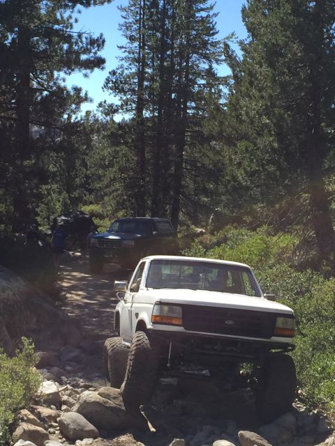 1989 Ford Bronco - Modified white pickup truck navigates a challenging rocky trail with another vehicle following.