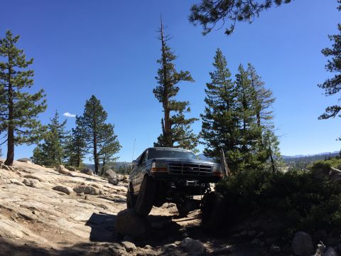1989 Ford Bronco - A dark-colored Ford Bronco is shown traversing a challenging, rocky off-road path under a bright blue sky.