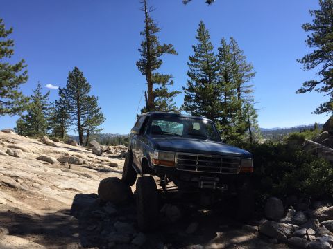 1989 Ford Bronco - A dark blue SUV navigates a rocky off-road trail under a clear blue sky.