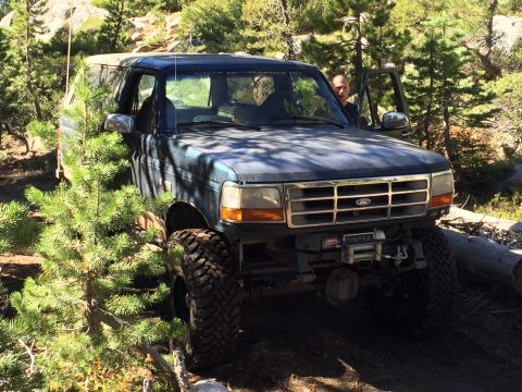 1989 Ford Bronco - A man stands by the open door of a dark blue 1992 Ford Bronco as it navigates a wooded trail.