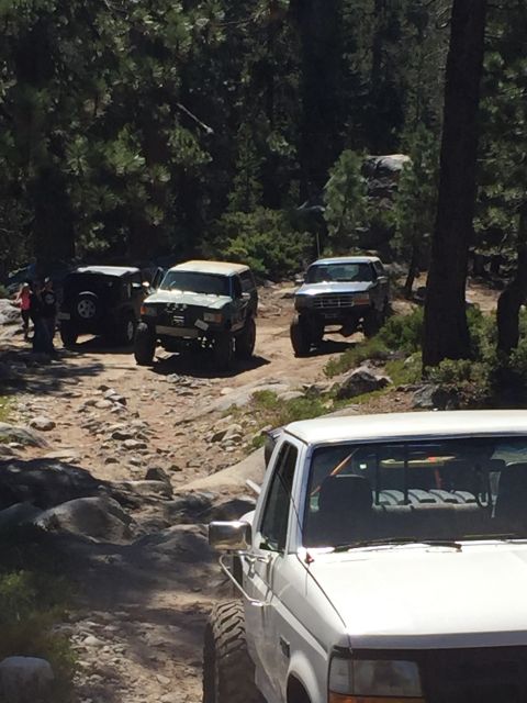 1989 Ford Bronco - Multiple off-road vehicles are seen on a dirt trail surrounded by trees and rocks.