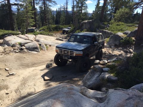 1989 Ford Bronco - A dark blue Ford Bronco navigates a rocky off-road trail with other vehicles visible in the distance.