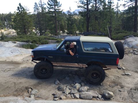 1989 Ford Bronco - A man drives a lifted Ford Bronco off-road through a rocky, wooded landscape.