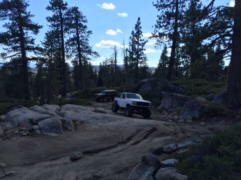1989 Ford Bronco - Two trucks navigate a challenging off-road path through a wooded area with large boulders.