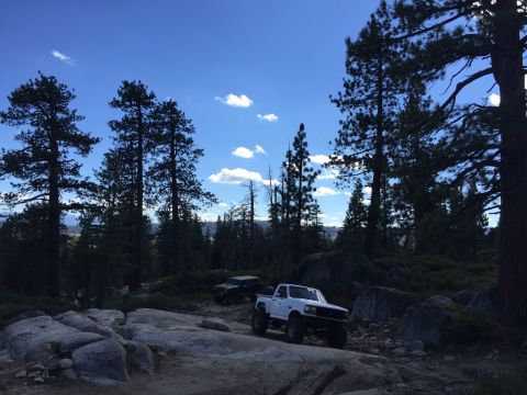 1989 Ford Bronco - Two off-road vehicles navigate a challenging rocky path amidst a forest landscape under a clear blue sky.