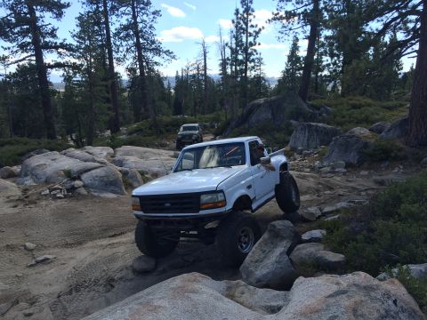 1989 Ford Bronco - A white Ford pickup truck is shown off-roading on a rocky trail with another vehicle in the background.