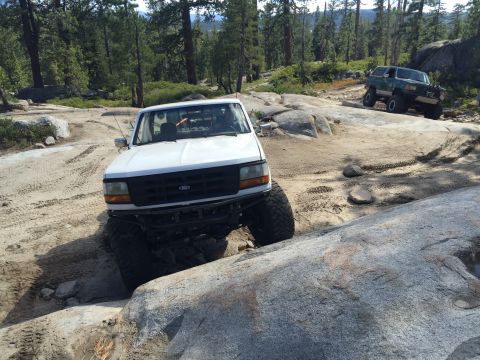 1989 Ford Bronco - A white Ford F-150 truck is shown navigating a challenging rocky off-road course, with another vehicle visible in the background.