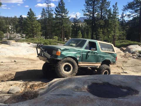 1989 Ford Bronco - A modified green Ford Bronco navigates a challenging off-road course amidst trees and rocks.