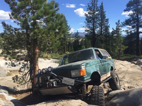1989 Ford Bronco - Modified green Ford Bronco tackling a challenging off-road trail with large rocks and trees.