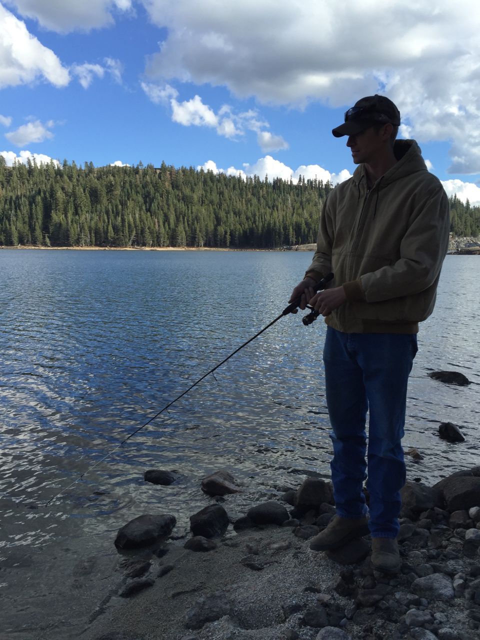 Man wearing a baseball cap and jacket fishes from the rocky shore of a lake with a tree-covered mountain range in the background