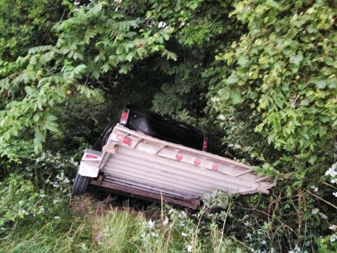 1992 Ford Bronco - A black pickup truck and trailer are off the road, surrounded by dense foliage.