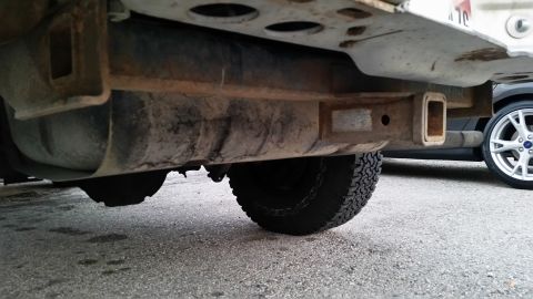 1981 Ford Bronco - The underside of a vehicle shows a rusty trailer hitch receiver mounted to the frame, with a large fuel tank visible above it. A tire with…