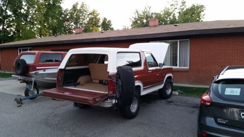 1981 Ford Bronco - A red and white Ford Bronco with its rear hatch and hood open is parked next to a red Bronco towing a small boat. A dark gray hatchback is…