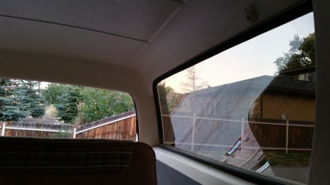 1981 Ford Bronco - View from the interior of a vehicle looking out the rear side window at a wooden fence and trees. The window reflects a house and a gravel…