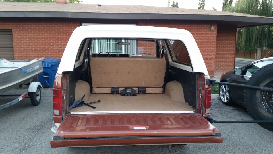 Rear view of a brown and white Ford Bronco with tailgate open, showing cargo area. Boat on trailer to the left.
