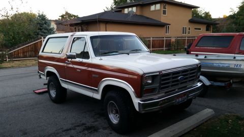 1981 Ford Bronco - A two-tone brown and white 1980s Ford Bronco is parked on an asphalt surface next to a boat on a trailer. The Bronco is viewed from a 3/4 f…