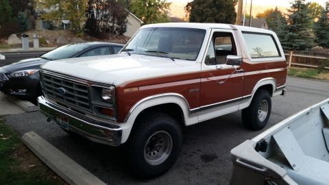 1981 Ford Bronco - A white and brown 1980s Ford Bronco is parked on a residential street next to a dark gray sedan. The Bronco is shown from a 3/4 front angle…