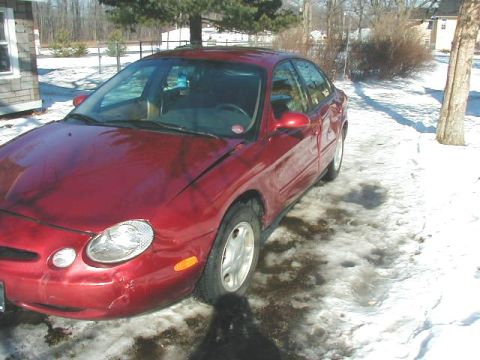 1997 Ford Taurus - Front three-quarter view of a red Ford Taurus sedan parked on snow-covered ground, showing a creased and dented driver's-side front fender…